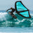 A windsurfer in a black wetsuit rides a turquoise and black windsurf board over a turquoise ocean wave under a clear blue sky.