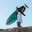 A person carrying a turquoise and black North Surfboard in a desert setting under a clear blue sky.