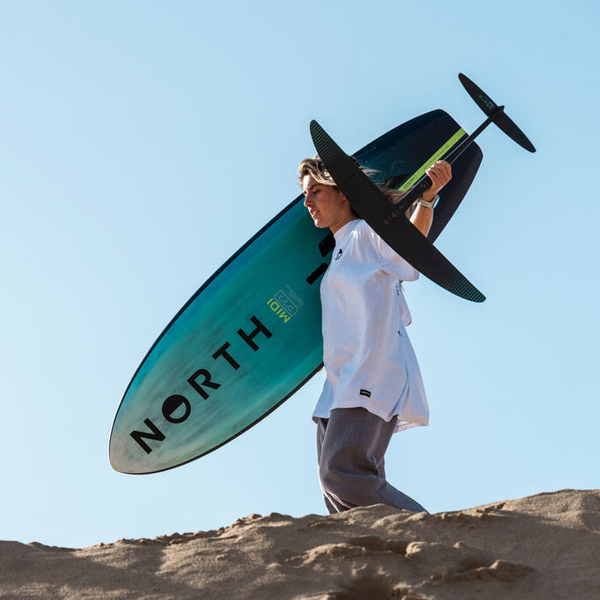 A person carrying a turquoise and black North Surfboard in a desert setting under a clear blue sky.